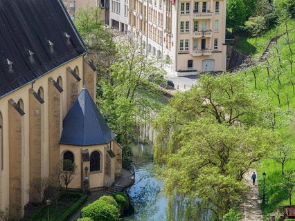 View of a church on the river bank, with neighbouring buildings and surrounded by lush greenery and