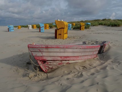 Old, half buried boat in front of colourful beach chairs on sandy beach, cloudy sky and dunes,