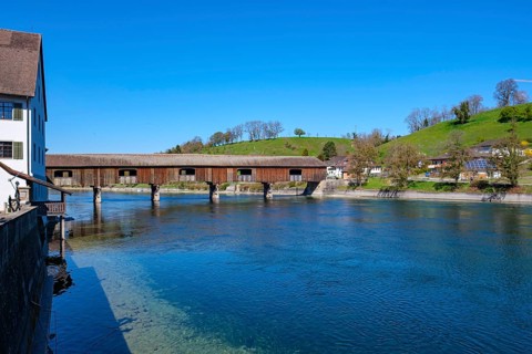 Wooden bridge over a river with old buildings in the background and a green meadow under a bright