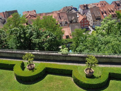 View of a picturesque garden with trees and trimmed hedges, in the background roofs and a lake, old