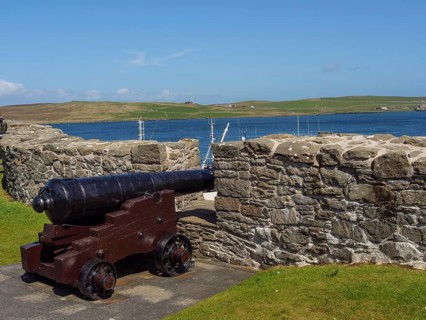 Old cannon standing in front of a stone wall with sea view and green hills under blue sky, old