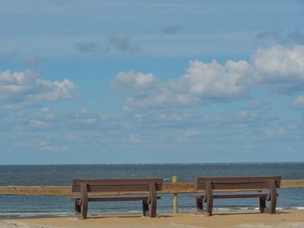 Two wooden benches overlooking the calm and wide sea under a cloudy sky, beach and dunes with grass