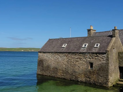 Small stone house on the shore of clear blue water with sweeping views of the coastline and blue