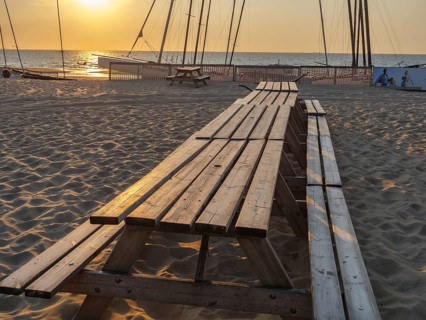 Long picnic tables on sandy beach at sunset, boats in the background, warm and relaxing atmosphere,