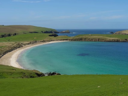 Calm bay with golden sandy beach and blue water, surrounded by green grassland and a clear sky,