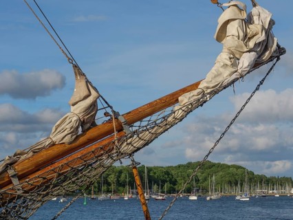 A wooden ship's bow with sail and rig in the foreground, colourful boats and wooded coast in the