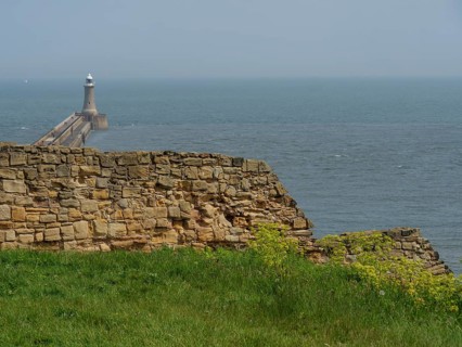 A lighthouse on a pier with old wall remains and green grass in the foreground, ruins and old stone