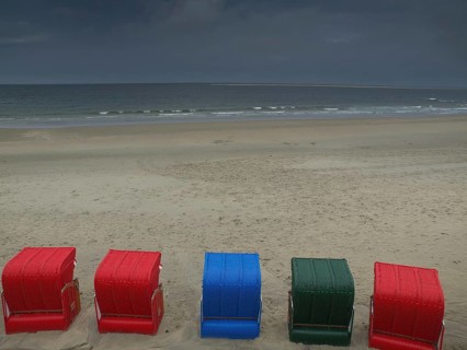 Five colourful beach chairs in red, blue and green stand on a quiet beach in front of a cloudy sky,