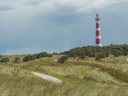 A red and white striped lighthouse in a grassy and wooded dune landscape under a cloudy sky, red