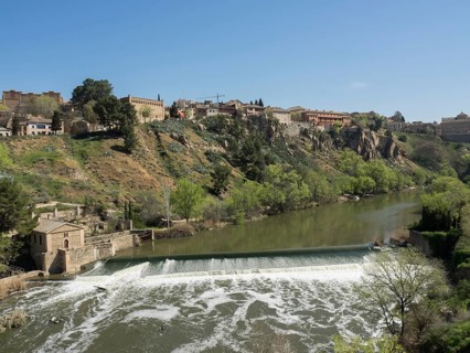 A city view on a hill with buildings, a river with a waterfall and green areas under a clear sky,