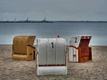 Three beach chairs on a sandy beach in front of the calm sea, ships on the horizon under a cloudy