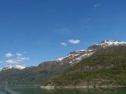 A clear day with snow-capped mountains under a blue sky with scattered clouds, bridge in a fjord