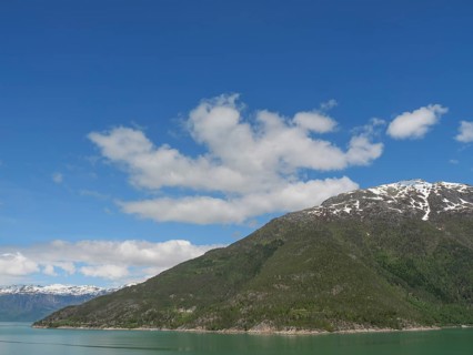 A mountain under a clear blue sky with some clouds, surrounded by green forests and a lake,