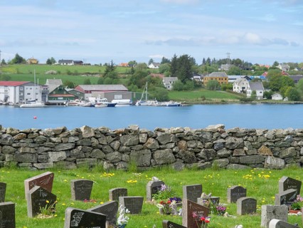 Cemetery with many gravestones, behind it a lake with boats and houses on the hill, under a