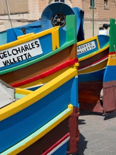 Several colourful boats on land with the inscriptions ANGEL CHARLES VALLETTA and ROCKY II VALLETTA,