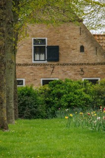 Brick house with black shutters and numbered facade, surrounded by a green meadow and flowers, old