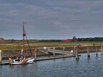 A sailing boat is moored at a pier, houses and people can be seen in the background, all under a