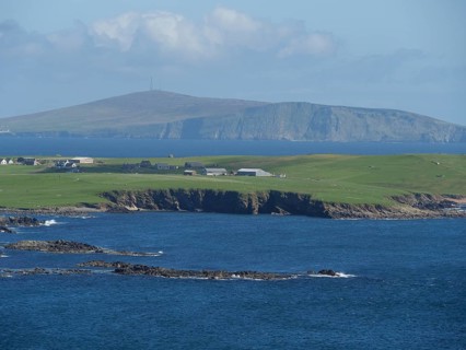 Green fields stretch to the jagged cliffs in front of the deep blue sea and a distant island, green