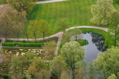 A green park with footpaths, a pond and trimmed hedges, surrounded by trees in spring, small park