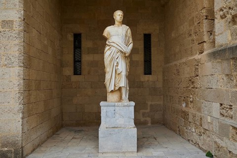 Ancient statue of a man in a long robe on a pedestal in front of a stone wall, torso from the 2nd