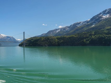 Turquoise lake surrounded by snow-capped mountains, a bridge spans in the background under a blue