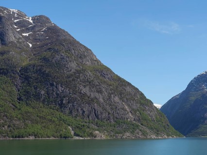 Steep rock faces covered with a forest reach down to a clear water and snow-capped mountain peaks