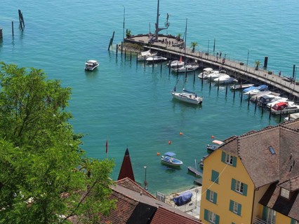 View of a harbour with boats at the pier, behind it houses and a calm blue lake, old buildings with