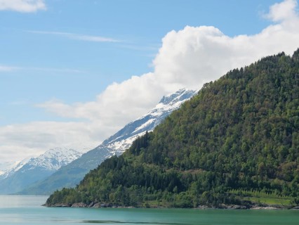 Green mountain slopes with snow-covered peaks and a lake under a slightly cloudy sky, greenish
