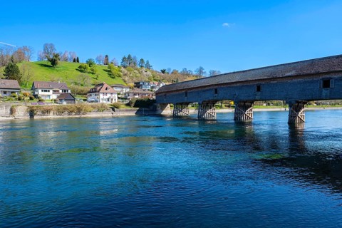 Massive wooden bridge over a clear river, surrounded by houses and green hills, under a blue sky,