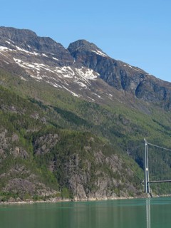 Snow-capped mountains with forested flanks, prominent bridge over turquoise lake under blue sky,