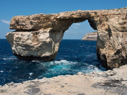 Natural rock arch stretches over the sea, surrounded by coastal cliffs under a blue sky, many