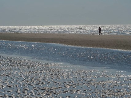 Single person walking along a wide beach with reflecting watercourse, glittering sea water at low