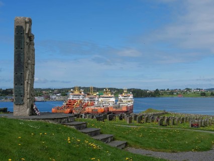 Stone memorial in a cemetery with a view of the harbour and several anchored ships, old stone