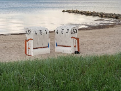 Two white beach chairs in front of a green meadow, sandy beach and calm sea in the background,