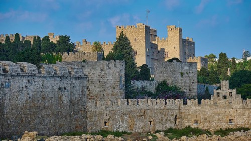 Ancient fortress walls with battlements, surrounded by lush greenery under a partly cloudy sky,