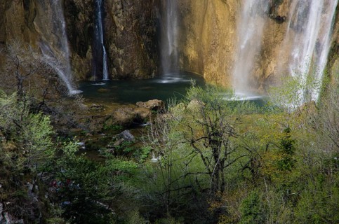 A waterfall cascades down between rocks, surrounded by green foliage and trees. Plitvice Lakes