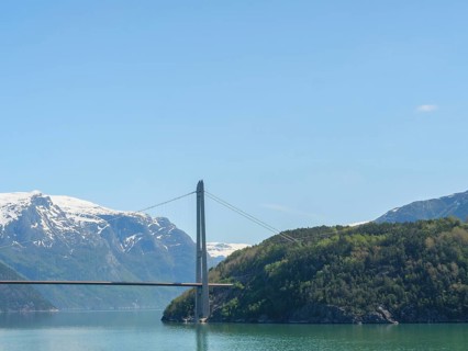Landscape with high bridge, snow-capped mountains and green wooded hills under a clear blue sky,