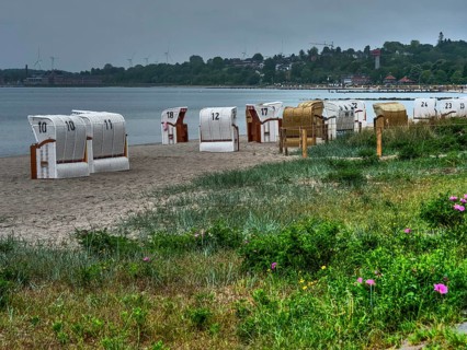 Beach chairs stand in a row on a sandy beach along the coast. The sky is cloudy, houses can be seen