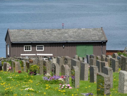 Wooden house behind a cemetery with many flowers on the shore of a lake, old stone church and many