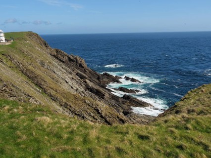 Steep cliffs with green grass and views of the azure sea and undulating coastline under a blue sky,