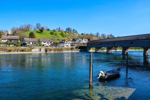 View of a calm river with a boat, a bridge and houses along the shore, embedded in a green hilly