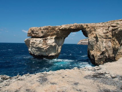 Rock arch over the sea, surrounded by cliffs, under a clear blue sky, many historic windmills by a