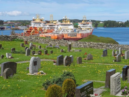 Cemetery with gravestones and three big orange ships by the sea, under a blue, slightly cloudy sky,