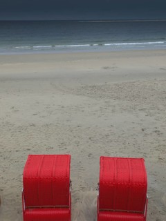 Two red beach chairs stand on an empty beach in front of a calm sea under a cloudy sky, beach