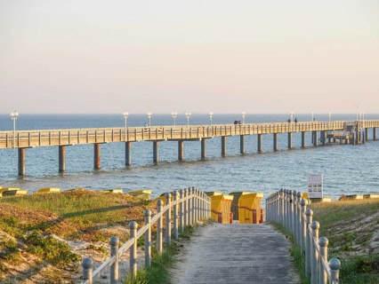 A long jetty leads into the sea, next to it beach chairs and a yellow sky at sunset, beach chairs
