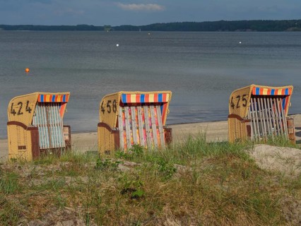 Three beach chairs with colourful stripes stand on a sandy beach in front of a calm sea and cloudy