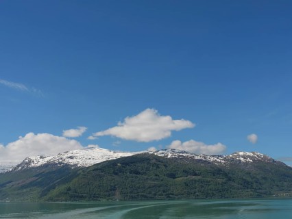 A mountain with snow peaks under a blue sky with clouds, in front of a calm lake, greenish