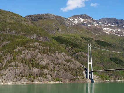 Concrete bridge stretching across a mountain landscape with dense forests and snow-capped peaks,
