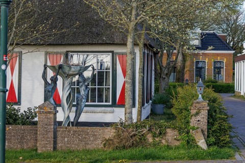 House with large windows and red curtains, in front of it stands a modern sculpture under trees,