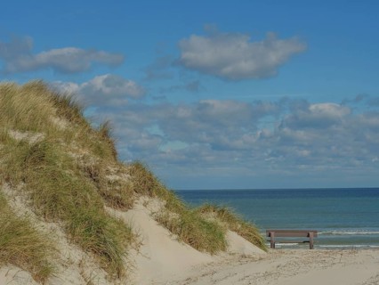 Sand dunes with grass, behind them the sea and a bench, above them a cloudy sky, dunes on an island